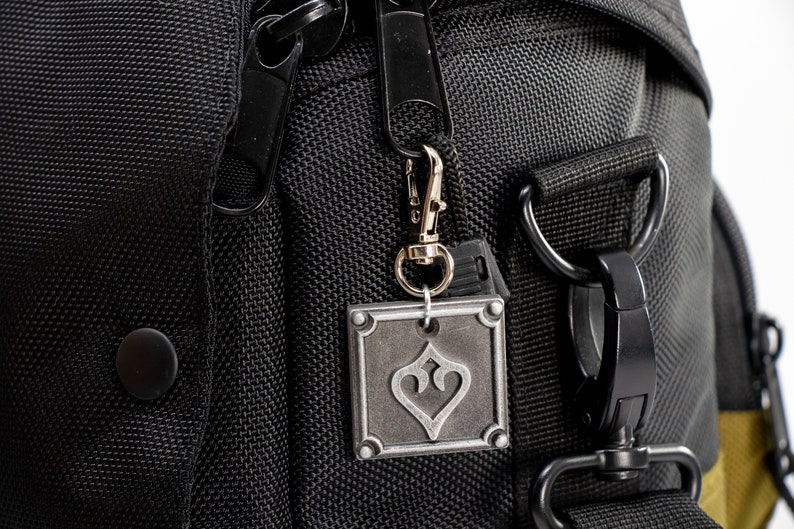 May include: Black zippered bag with a silver metal charm featuring a stylized heart design. The bag has a black strap with a metal buckle.