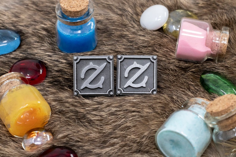 Op de afbeelding: Twee zilveren metalen pinnen met een gestileerde letter Z op een bruin, pluche oppervlak. De pinnen zijn omringd door kleine glazen flesjes gevuld met gekleurd zand.