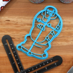 May include: A blue plastic cookie cutter shaped like a cartoon character with a large head and a body with details. The cookie cutter is on a wooden surface.