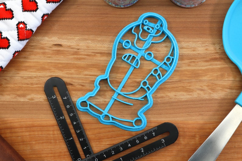 May include: A blue plastic cookie cutter shaped like a cartoon character with a large head and a body with details. The cookie cutter is on a wooden surface.