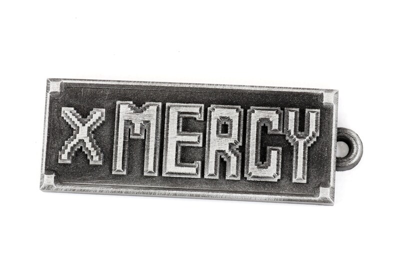 May include: A silver metal keychain with the text "X MERCY" in a pixelated font.