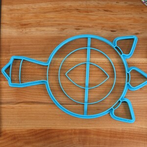 May include: A blue plastic cookie cutter shaped like a turtle with a circle in the center and a smaller circle inside the larger circle. The turtle has a long neck and three points on its shell.