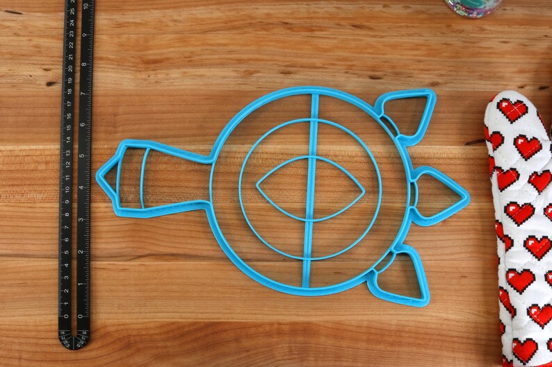 May include: A blue plastic cookie cutter shaped like a turtle with a circle in the center and a smaller circle inside the larger circle. The turtle has a long neck and three points on its shell.