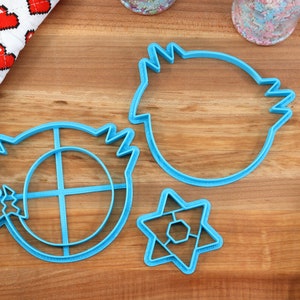 May include: Three blue plastic cookie cutters on a wooden surface. The cutters are shaped like a star, a circle with a cross inside, and a round shape with a jagged edge.