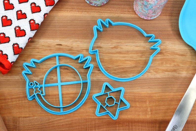 May include: Three blue plastic cookie cutters on a wooden surface. The cutters are shaped like a star, a circle with a cross inside, and a round shape with a jagged edge.