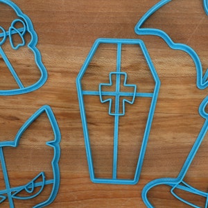 May include: A set of five blue plastic cookie cutters shaped like a witch's hat, a bat, a coffin, a cat, and a ghost. The cutters are on a wooden surface.
