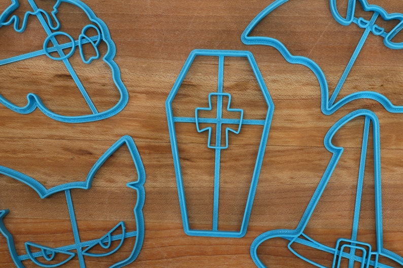 May include: A set of five blue plastic cookie cutters shaped like a witch's hat, a bat, a coffin, a cat, and a ghost. The cutters are on a wooden surface.