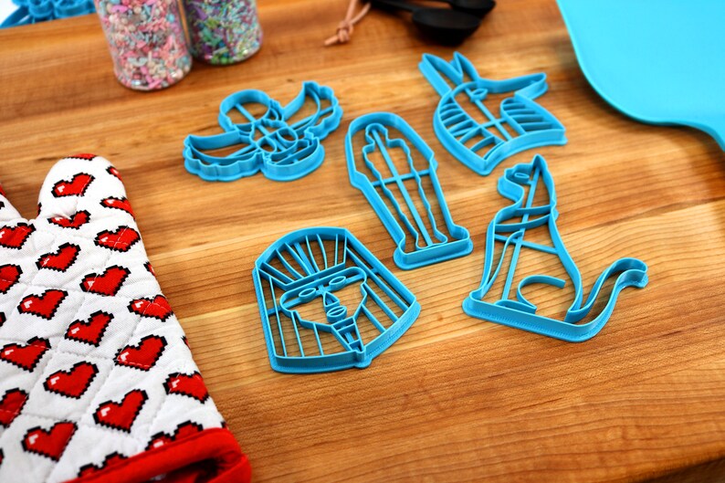 May include: A set of six blue plastic cookie cutters shaped like Egyptian symbols, including a pharaoh's head, a scarab beetle, an ankh, a cat, a pyramid, and a sarcophagus.