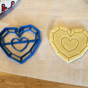 May include: A blue plastic cookie cutter shaped like a heart with a smaller heart inside. A heart-shaped cookie is on a white parchment paper.