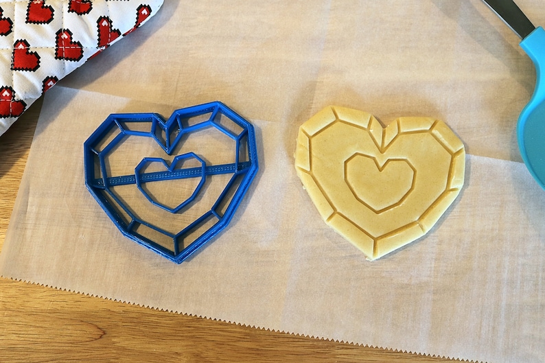 May include: A blue plastic cookie cutter shaped like a heart with a smaller heart inside. A heart-shaped cookie is on a white parchment paper.