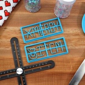 May include: Two blue plastic cookie cutters on a wooden surface. The cutters are shaped to cut out the words "Merry" and "Item".