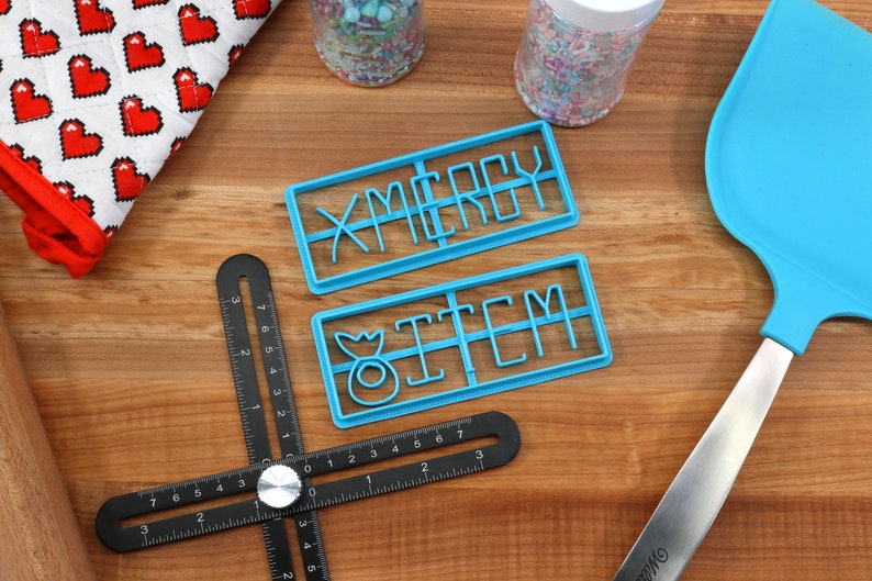 May include: Two blue plastic cookie cutters on a wooden surface. The cutters are shaped to cut out the words "Merry" and "Item".