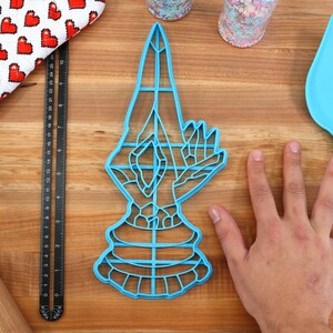 May include: A blue plastic cookie cutter shaped like a hand in a prayer position. The cookie cutter is about 7 inches tall and is sitting on a wooden surface.