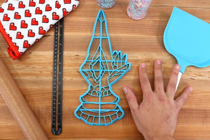 May include: A blue plastic cookie cutter shaped like a hand in a prayer position. The cookie cutter is about 7 inches tall and is sitting on a wooden surface.