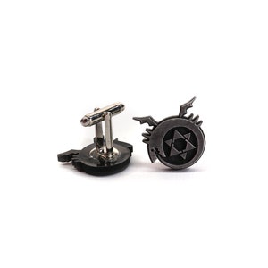 May include: A pair of cufflinks featuring the black and silver symbol of the Fullmetal Alchemist, a popular anime series. The symbol is a circle with a six-pointed star inside, and a winged creature on the outside.