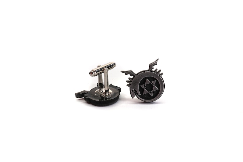 May include: A pair of cufflinks featuring the black and silver symbol of the Fullmetal Alchemist, a popular anime series. The symbol is a circle with a six-pointed star inside, and a winged creature on the outside.