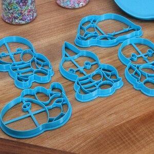 May include: Five blue plastic cookie cutters shaped like cartoon characters. The cutters are on a wooden surface.