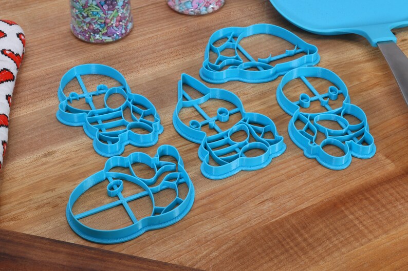 May include: Five blue plastic cookie cutters shaped like cartoon characters. The cutters are on a wooden surface.