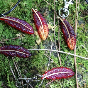 May include: Five red and gold feather charms with a metallic sheen. The charms are on a black cord, a silver chain, and a silver key ring. The charms are on a bed of green moss.