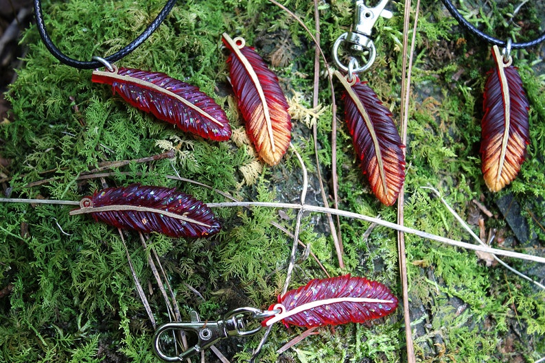 May include: Five red and gold feather charms with a metallic sheen. The charms are on a black cord, a silver chain, and a silver key ring. The charms are on a bed of green moss.