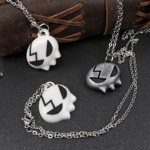 May include: Two silver chains with pendants. One pendant is white with black details and the other is silver with black details. Both pendants are shaped like a face with a lightning bolt design.