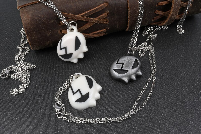 May include: Two silver chains with pendants. One pendant is white with black details and the other is silver with black details. Both pendants are shaped like a face with a lightning bolt design.