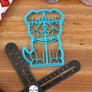 May include: A blue plastic cookie cutter shaped like a dog. The dog has a happy expression with a tongue sticking out. The cookie cutter is on a wooden surface.