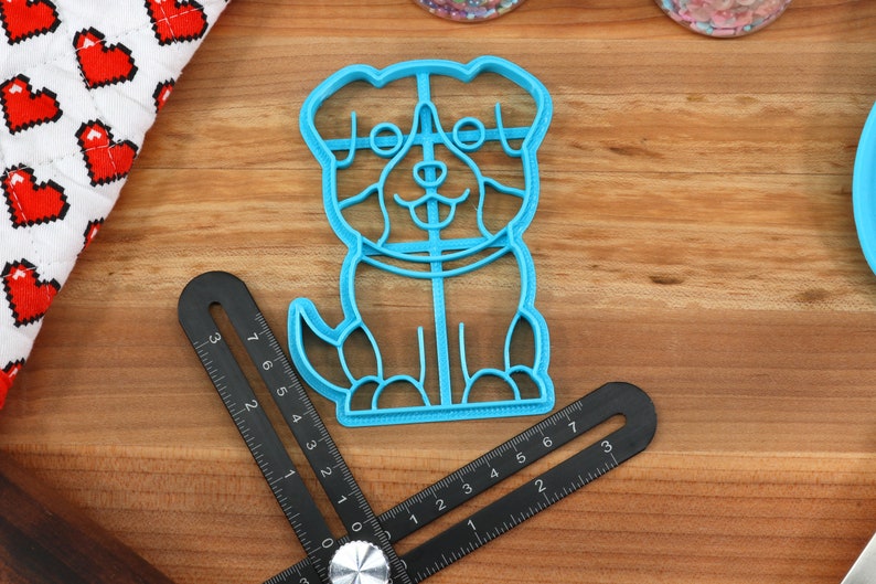 May include: A blue plastic cookie cutter shaped like a dog. The dog has a happy expression with a tongue sticking out. The cookie cutter is on a wooden surface.