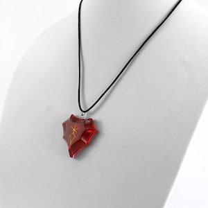 May include: A red heart-shaped pendant with a gold design hangs from a black cord necklace.