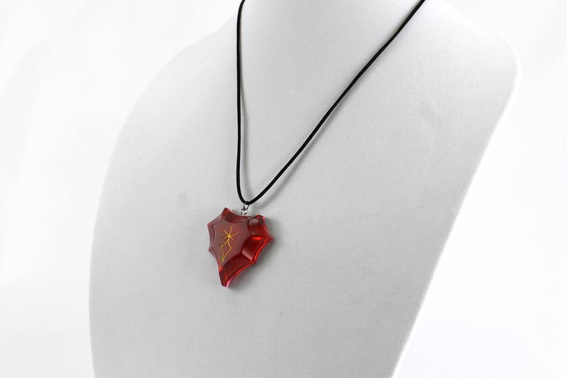 May include: A red heart-shaped pendant with a gold design hangs from a black cord necklace.