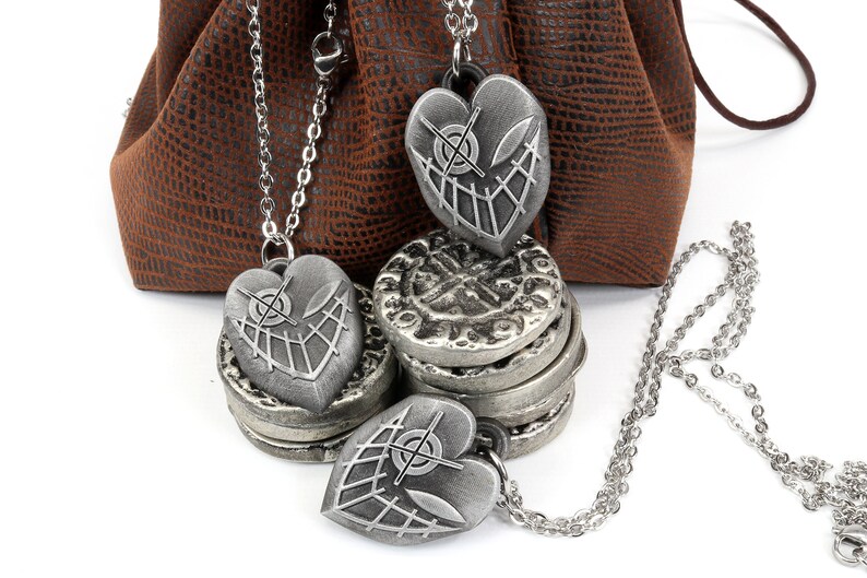 May include: Three silver heart-shaped pendants with a crosshatch design and a small circle in the center. The pendants are on chains and are hanging from a brown leather pouch.