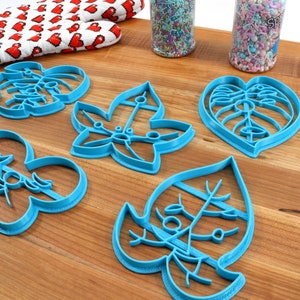 May include: Five blue plastic cookie cutters shaped like leaves. The cutters are on a wooden surface.