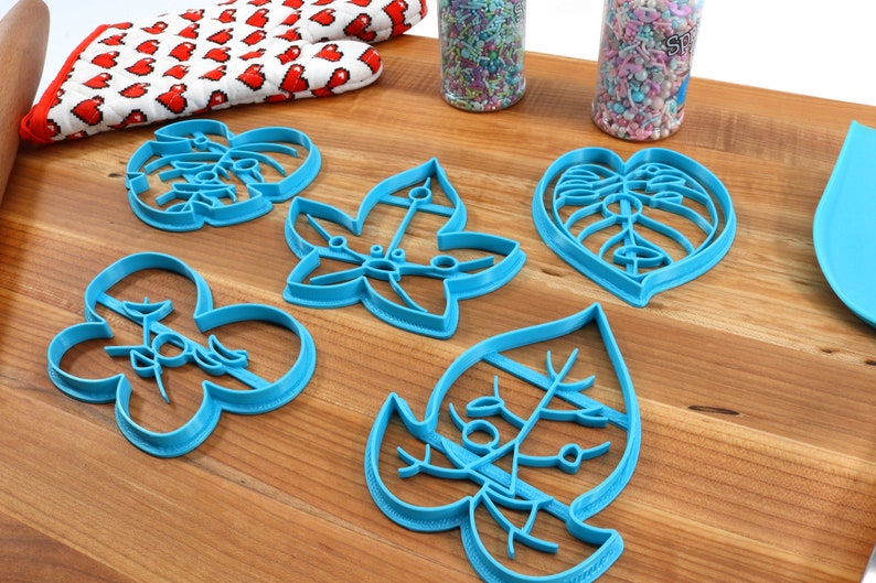 May include: Five blue plastic cookie cutters shaped like leaves. The cutters are on a wooden surface.