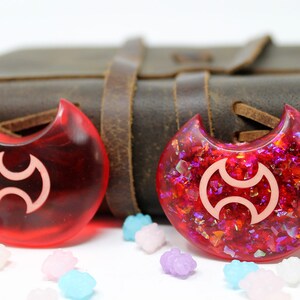 May include: Two red crescent moon-shaped resin charms with a white symbol in the center. One charm is clear and the other has red and iridescent glitter embedded in the resin.