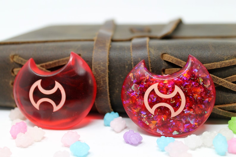May include: Two red crescent moon-shaped resin charms with a white symbol in the center. One charm is clear and the other has red and iridescent glitter embedded in the resin.