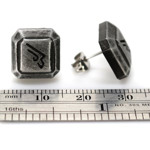 May include: A pair of silver-toned metal earrings with a geometric design. Each earring features a square-shaped piece with a stylized symbol inside. The earrings are shown next to a ruler, measuring approximately 1 cm in width.