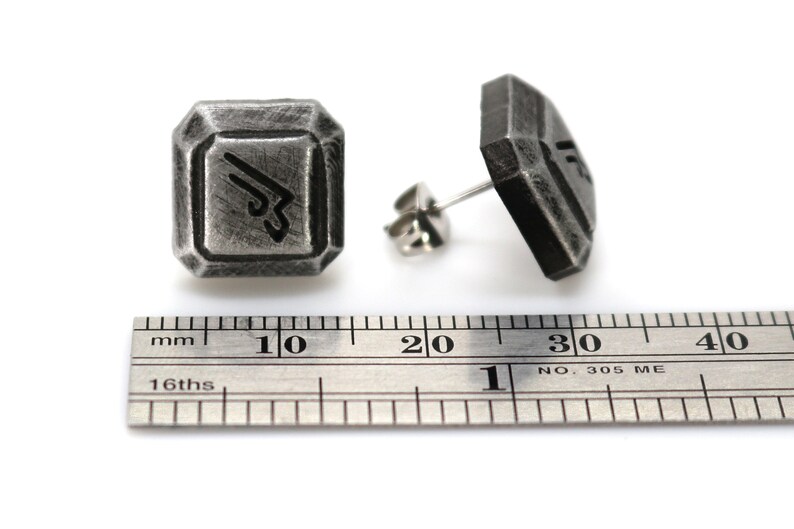May include: A pair of silver-toned metal earrings with a geometric design. Each earring features a square-shaped piece with a stylized symbol inside. The earrings are shown next to a ruler, measuring approximately 1 cm in width.