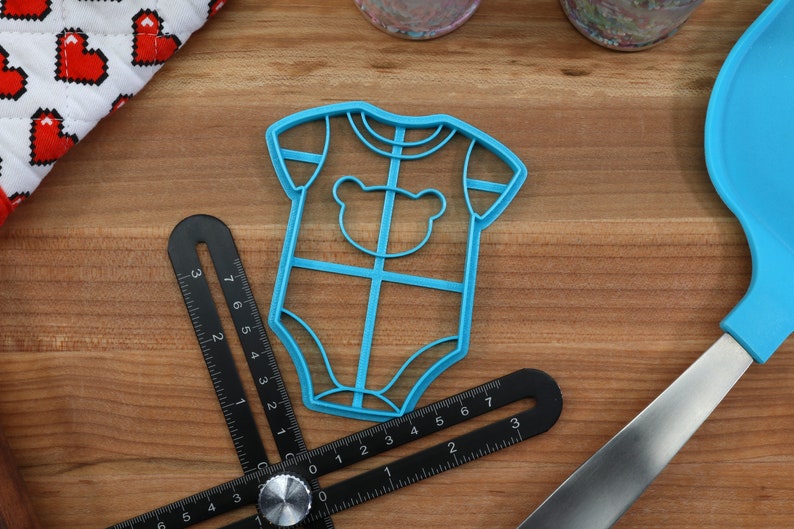 May include: A blue plastic cookie cutter shaped like a baby onesie with a bear face on the front.
