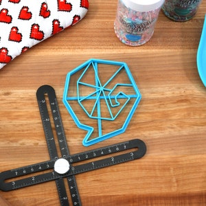May include: A blue plastic cookie cutter shaped like a spiral with a speech bubble. The cutter is on a wooden surface with a black ruler and a blue spatula.