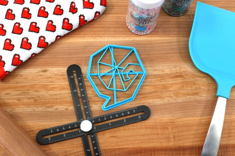 May include: A blue plastic cookie cutter shaped like a spiral with a speech bubble. The cutter is on a wooden surface with a black ruler and a blue spatula.