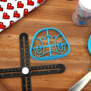 May include: A blue plastic cookie cutter shaped like a cartoon penguin face. The cutter has a grid pattern inside the face and is on a wooden surface.