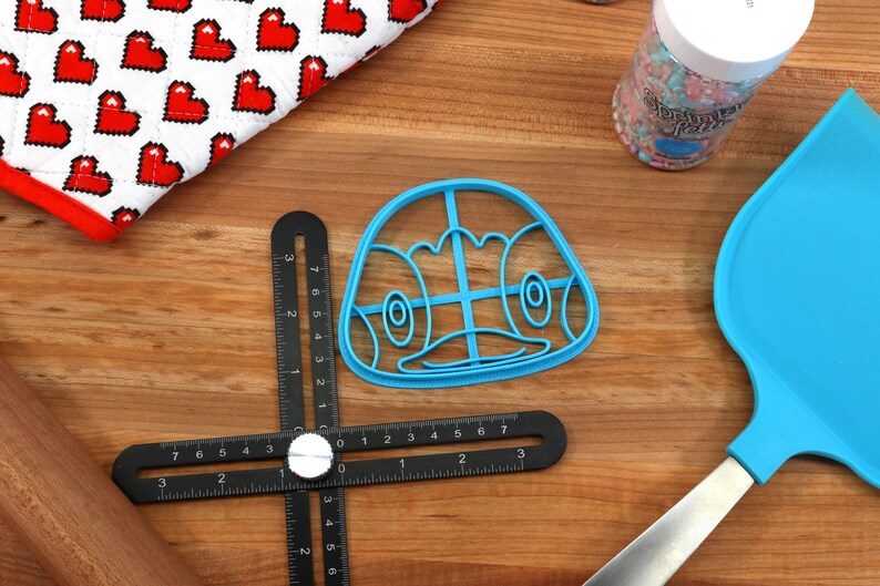 May include: A blue plastic cookie cutter shaped like a cartoon penguin face. The cutter has a grid pattern inside the face and is on a wooden surface.
