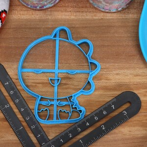 May include: A blue plastic cookie cutter shaped like a cartoon character with a smiling face and a spiky head. The cutter is on a wooden surface.