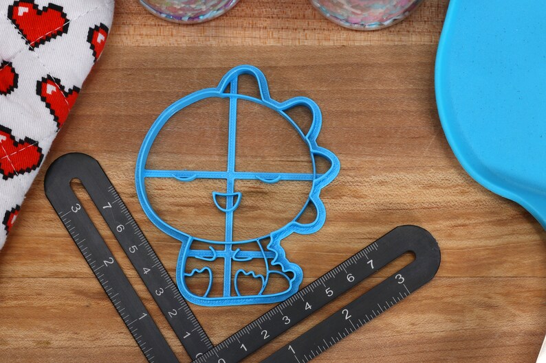 May include: A blue plastic cookie cutter shaped like a cartoon character with a smiling face and a spiky head. The cutter is on a wooden surface.