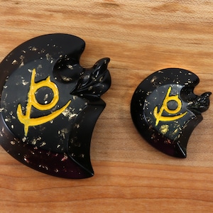 May include: Two black and gold decorative pieces with a stylized symbol in gold. The larger piece is shaped like a crescent moon and the smaller piece is round.