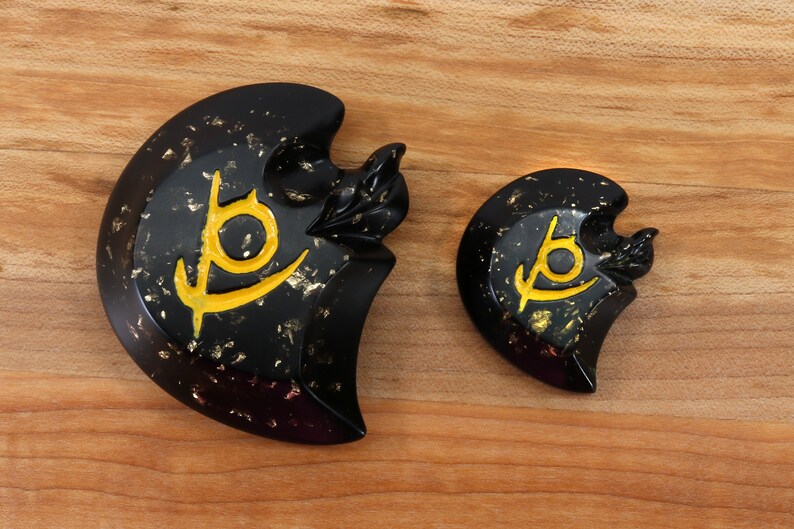 May include: Two black and gold decorative pieces with a stylized symbol in gold. The larger piece is shaped like a crescent moon and the smaller piece is round.