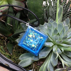 May include: A square blue resin pendant with iridescent glitter and a white design, hanging on a black cord. The pendant is displayed on a bed of green succulents.