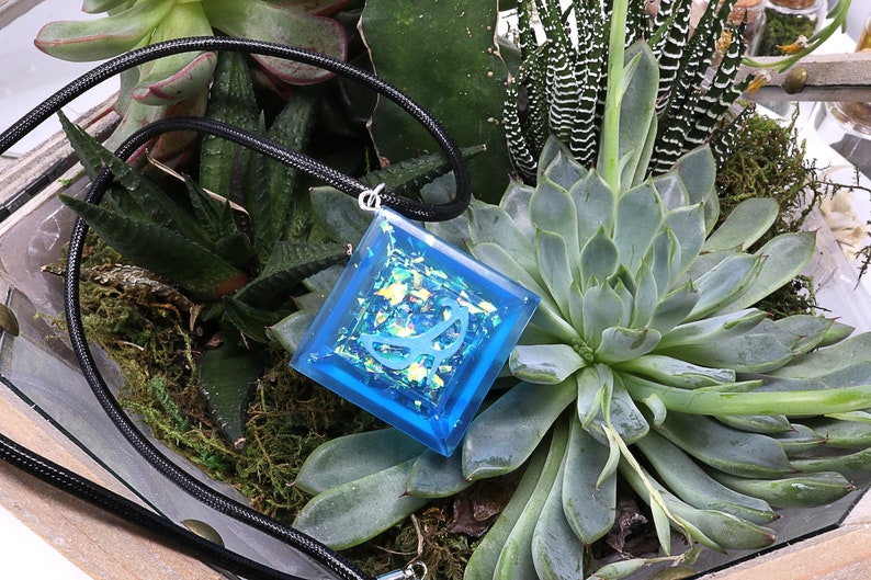 May include: A square blue resin pendant with iridescent glitter and a white design, hanging on a black cord. The pendant is displayed on a bed of green succulents.