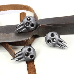 Op de afbeelding: Drie zilveren schedelvormige metalen bedels met elk drie gaten en vijf spikes. De bedels liggen op een bruin leren band en een roestig metalen mes.