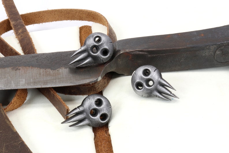 Op de afbeelding: Drie zilveren schedelvormige metalen bedels met elk drie gaten en vijf spikes. De bedels liggen op een bruin leren band en een roestig metalen mes.